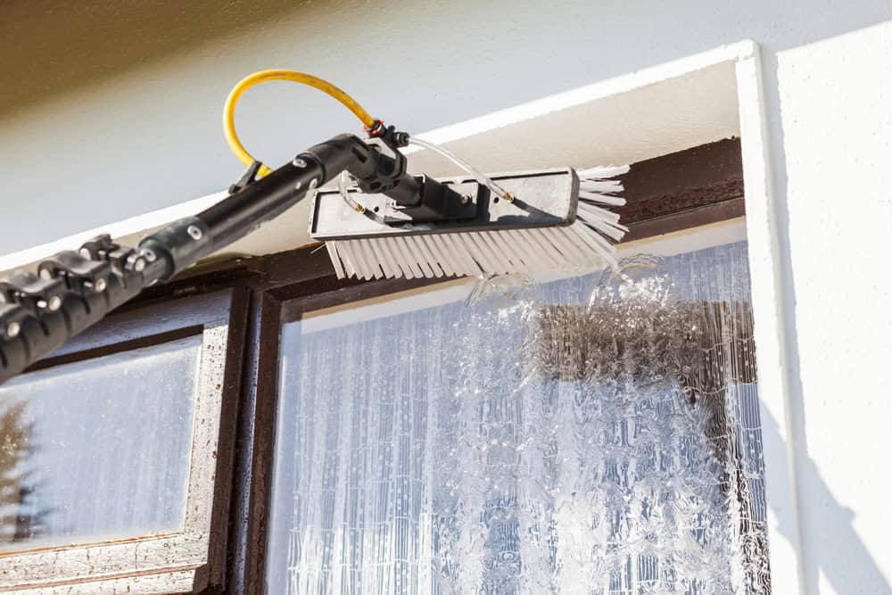 A close-up of a window being cleaned with a brush attached to a long pole, spraying water onto the glass and frame—showcasing expert pressure washing in Montgomery County, MD.