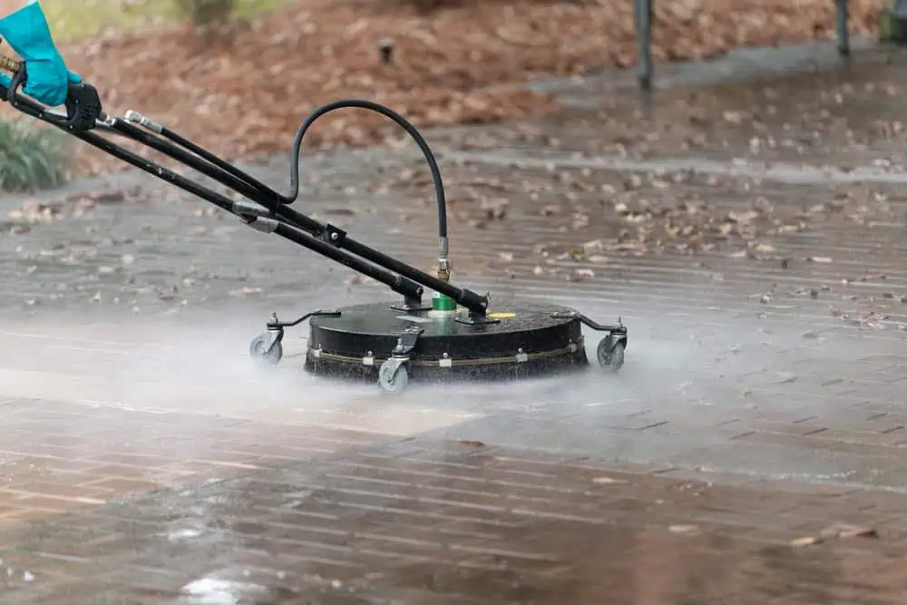 A person uses a circular surface cleaner attachment connected to a pressure washer to clean outdoor brick pavers in Montgomery County, MD. This pressure washing lifts dirt and grime, with water spraying outward to reveal a cleaner path.