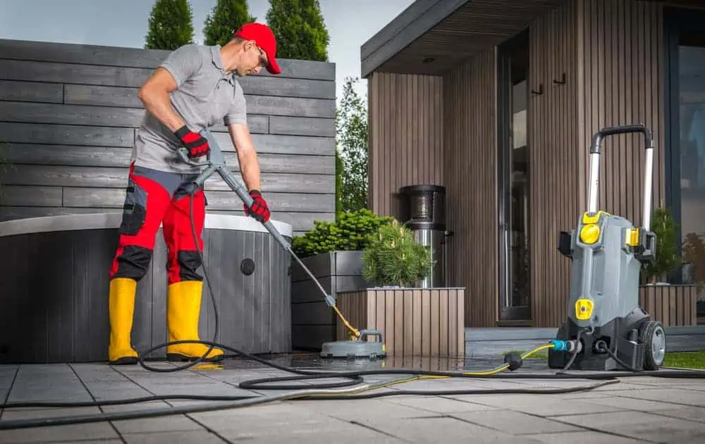 A man wearing yellow boots, red gloves, and a red cap is pressure washing a patio in Montgomery County, MD. A modern wooden house and potted plants are in the background, with power washer equipment visible in the foreground.