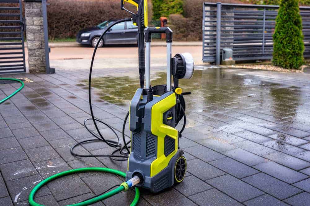 A yellow and gray pressure washer with a green hose attached stands on a wet, dark-tiled driveway—perfect for pressure washing Montgomery County, MD homes. A car, trees, and fencing can be seen in the background.